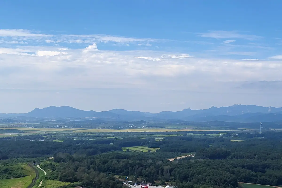 Een weids uitzicht naar het noorden vanaf Dora Observatory over groen bos en landbouwgrond richting een rij blauwe heuvels, met rechts een vlaggenmast en daarboven een grote bewolkte lucht