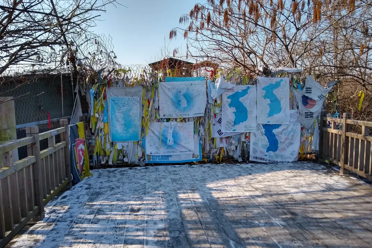 Linten, met de hand getekende kaarten van Korea en vlaggen vastgebonden aan een draadhek naast een houten wandelpad bij de Vrijheidsbrug, met kale bomen en een blauwe lucht op de achtergrond